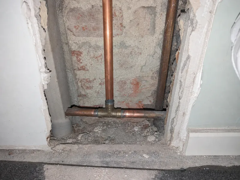 De foto bij bon MS-0148. Een opengebroken wand met roze baksteen erachter, twee koperen leidingen met een T-stuk, en gruis op de vloer ervoor.