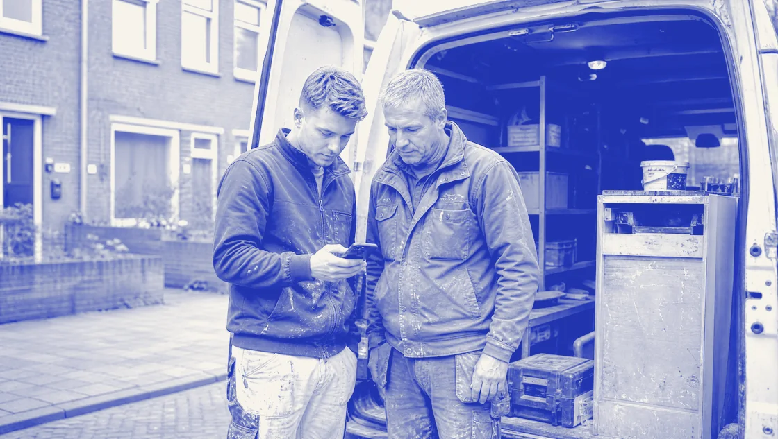 Twee vakmensen in werkkleding kijken samen naar een telefoon bij de open achterdeuren van een bedrijfsbus.